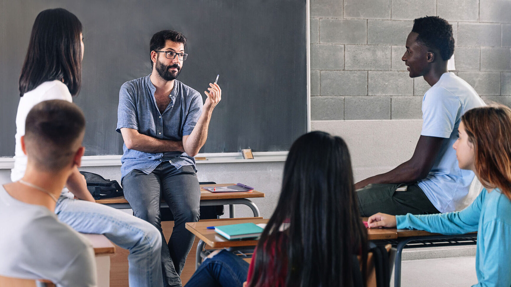 A young bearded teacher with glasses sits casually on his desk as he converses with teenage students after class. 