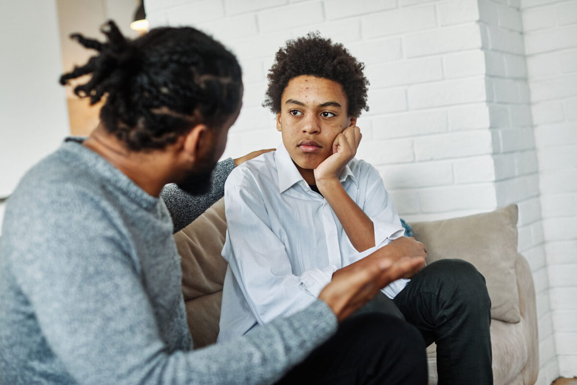 Portrait of a teenage boy listening to his father while sitting on the couch. The father has a comforting hand on the boy's shoulder.