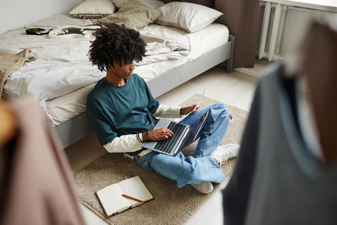 High angle portrait of teenage African-American student sitting on floor at home with laptop, studying