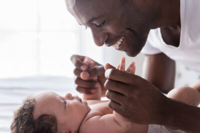 Side view of happy young African man playing with his little baby and smiling while lying in bed